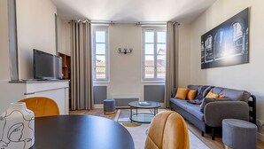 A modern living room with a sofa, a coffee table, and a TV on a stand. There are two windows with curtains, a painting on the wall, and a small table with a vase. - St Aubin - Gare & Capitole 10min - Hypercentre (Toulouse)