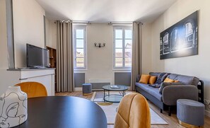 Moderni olohuone, jossa on sohva, sohvapöytä ja jalustalla oleva televisio. Huoneessa on kaksi ikkunaa, joissa on verhot, seinällä taulu ja pieni pöytä, jonka päällä on maljakko.