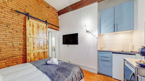 A bedroom with a bed, a wooden wardrobe, a television, and a kitchenette with blue cabinets and a sink.