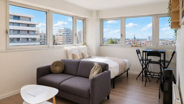 Ein modernes Apartment mit grauem Sofa, kleinem weißen Tisch, einem Bett und einem Essbereich. Große Fenster bieten Blick auf die Stadtlandschaft.