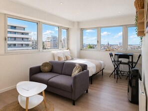 A modern apartment with a grey sofa, a small white table, a bed, and a dining area. Large windows offer views of the cityscape.