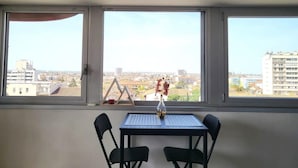 Ein Zimmer mit Tisch und zwei Stühlen, einer Vase mit Blumen und Blick auf Gebäude durch große Fenster.