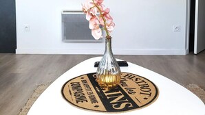 Ein modernes Wohnzimmer mit einem weißen Tisch, einer Vase mit rosa Blumen, einem gerahmten abstrakten Kunstwerk und einer kleinen Ablage mit Dekorationselementen.