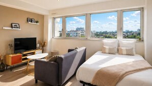 Ein modernes Hotelzimmer mit Bett, Sofa, Fernseher und großen Fenstern mit Blick auf die Stadt.