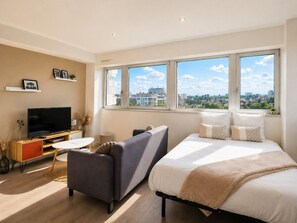 Een moderne hotelkamer met een bed, bank, televisie en grote ramen met uitzicht op de stad.