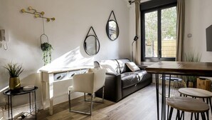 A modern living room with a black leather sofa, a white coffee table, and a dining area with stools. There are two wall-mounted mirrors and a hanging plant. - Parga 2 · Terrasse /Clim - Capitole - Bord Garonne (Toulouse)