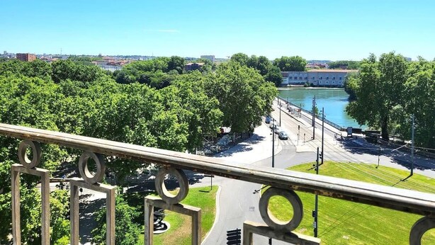 Vista del río desde un balcón con barandilla, árboles y un edificio distante al otro lado del agua.