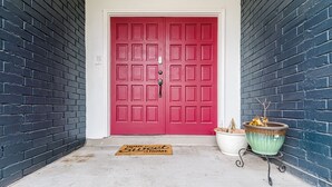 A red door with a white frame, flanked by dark brick walls, a white door mat, and a potted plant on a metal stand. - Cute home with a Backyard pool! (San Antonio)