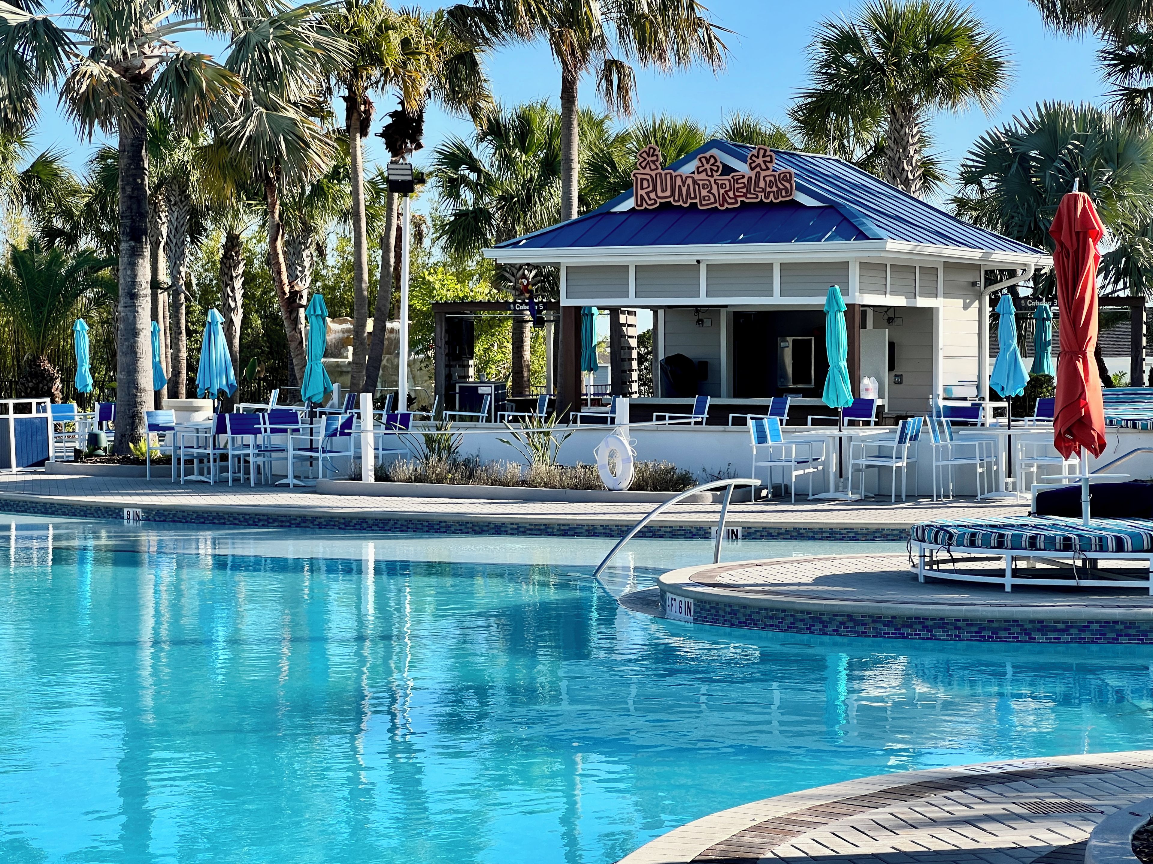 A pool area with lounge chairs and umbrellas, a pool with a blue edge, and a building with a sign that reads 'RUMBRELAS'.