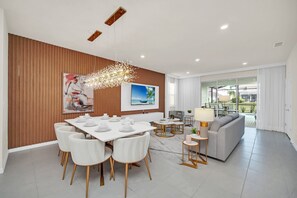 A modern dining area with a white table set for four, surrounded by white chairs. There is a large painting on the wall, a ceiling light fixture, and a view into a living area with a sofa and a table. - Avatar Game Room • Harry Potter & Princess Themed • Private Pool • Near Disney (Davenport)
