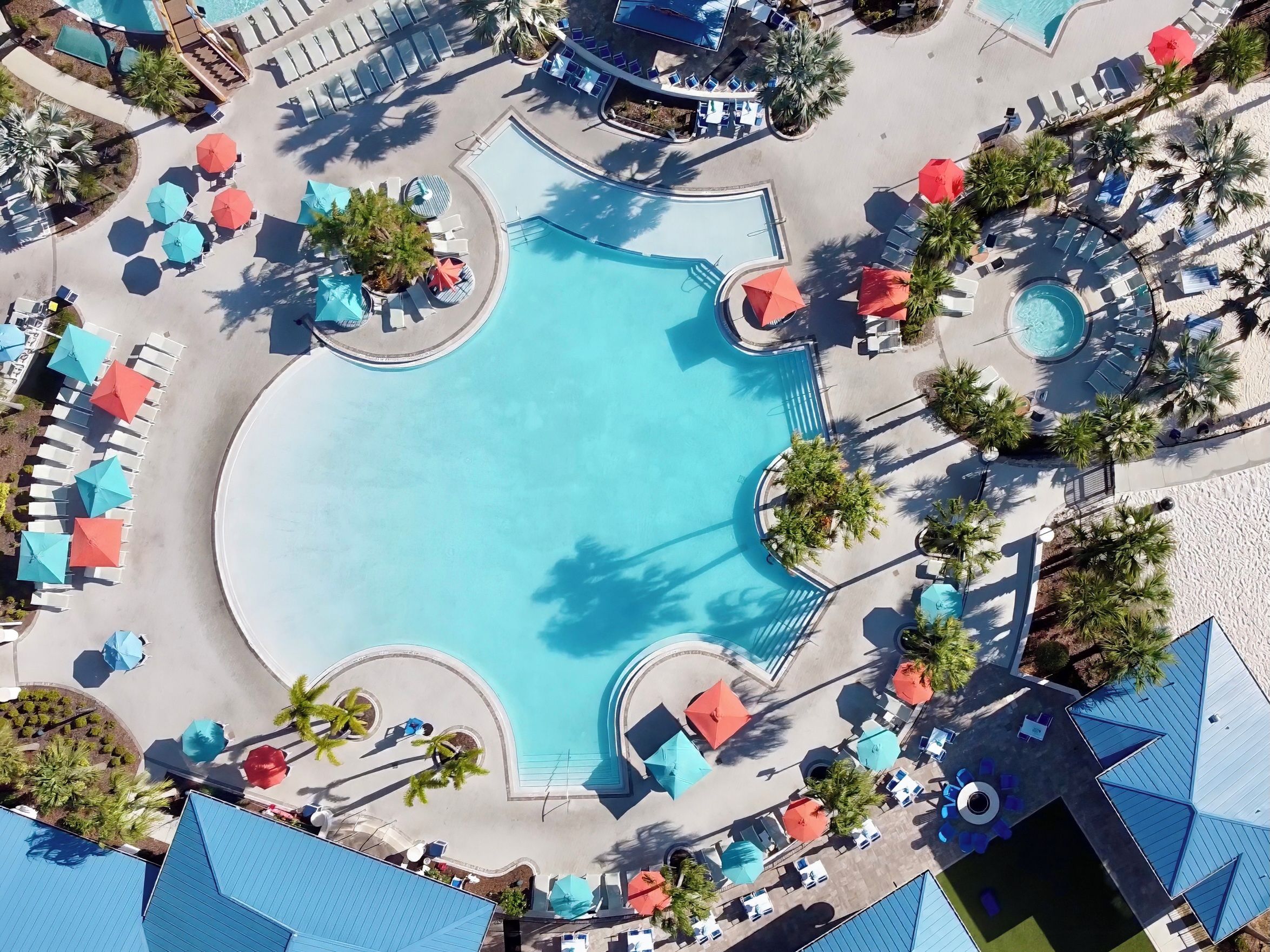 An aerial view of a resort with a central swimming pool, surrounded by lounge areas with umbrellas and sun loungers.