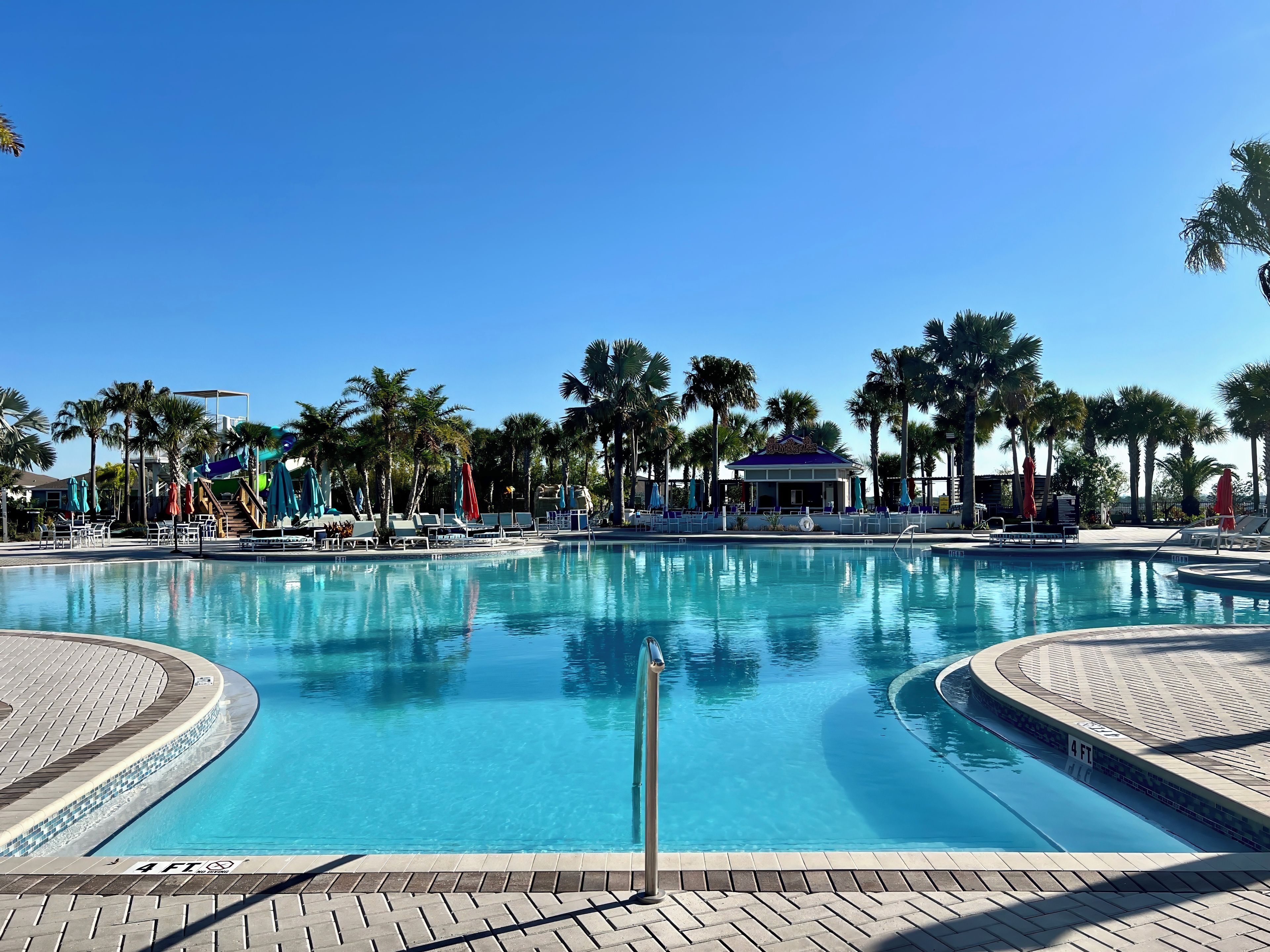 A clear blue swimming pool with a 4 ft depth marker, surrounded by palm trees and lounge chairs.