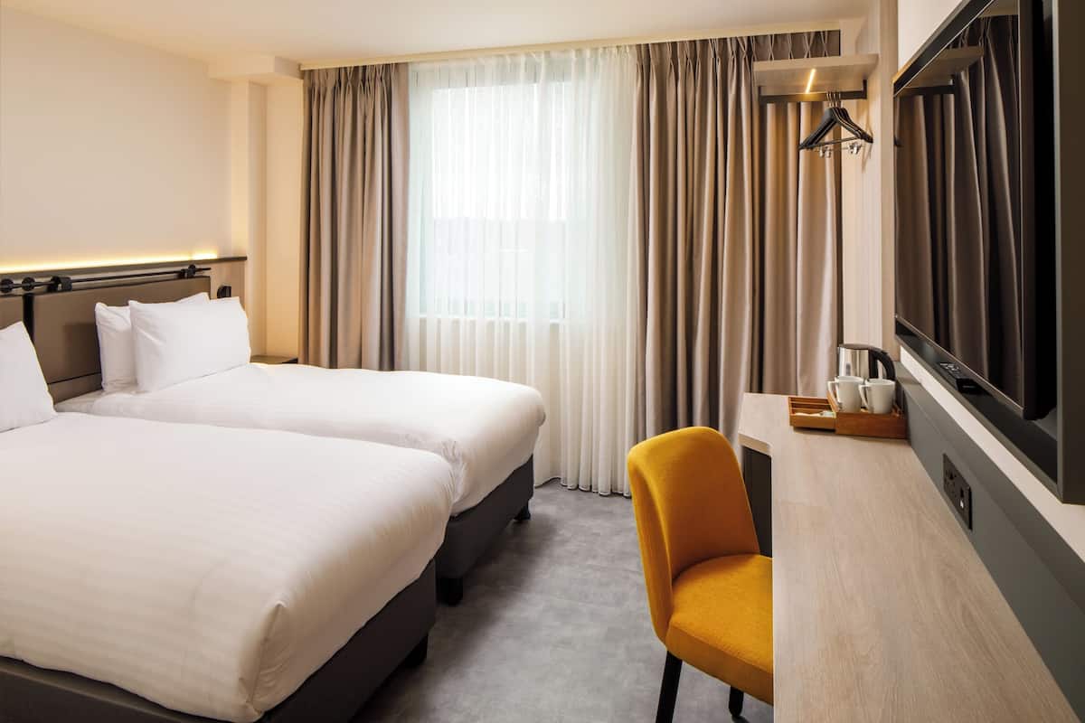 A hotel room with two beds, a desk with a TV, a chair, and a window with curtains.