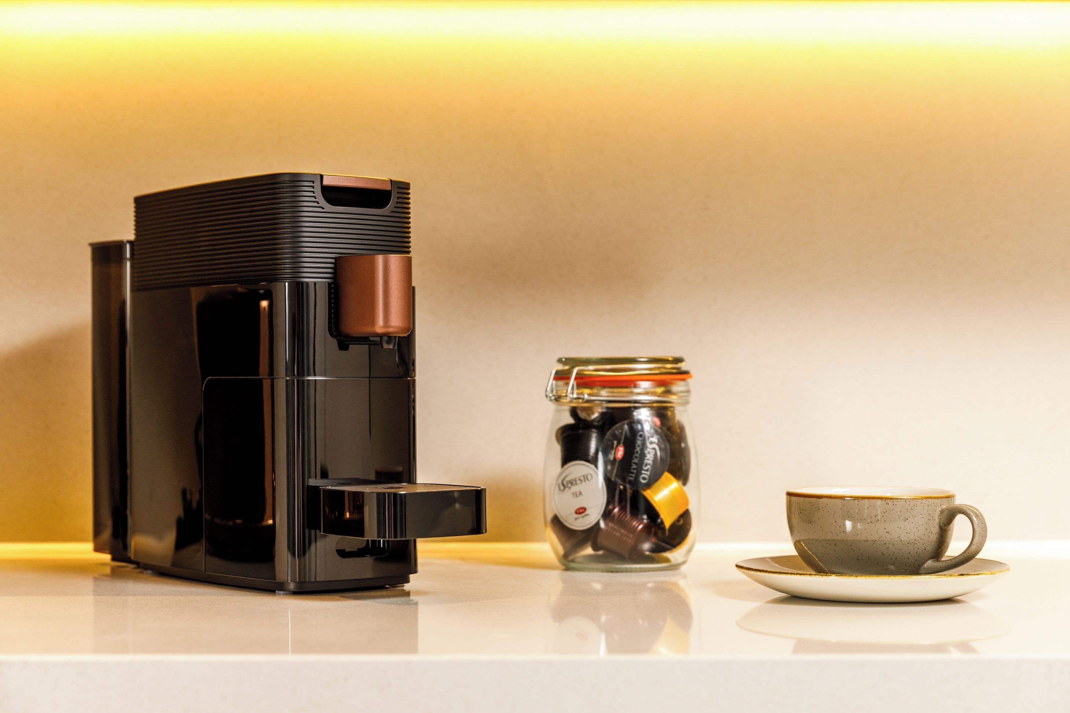 Eine Kaffeemaschine, ein Behälter mit Kaffeekapseln und eine Tasse auf einer Arbeitsplatte.