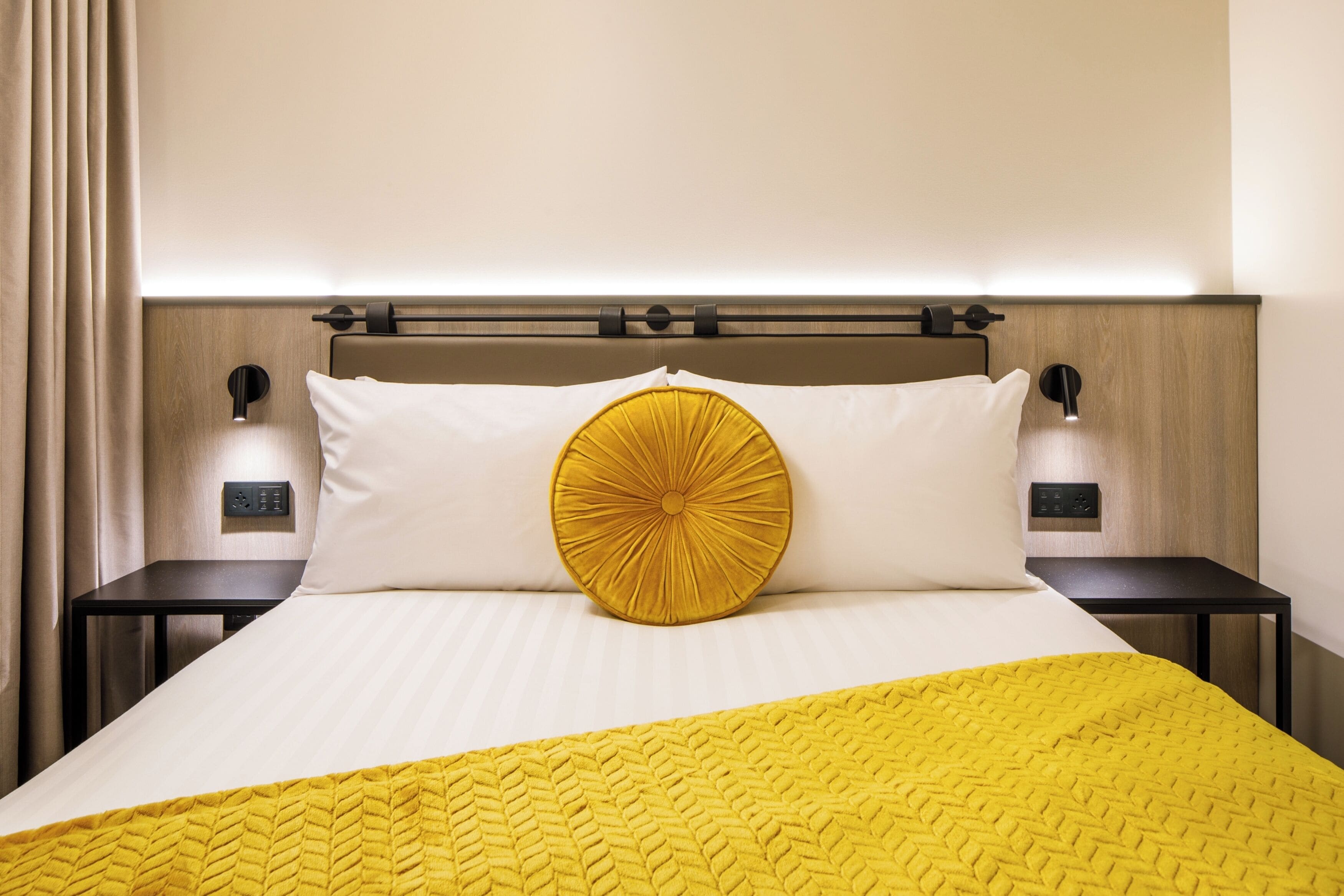 A hotel room with a bed, a nightstand, a wall-mounted light, and a control panel.