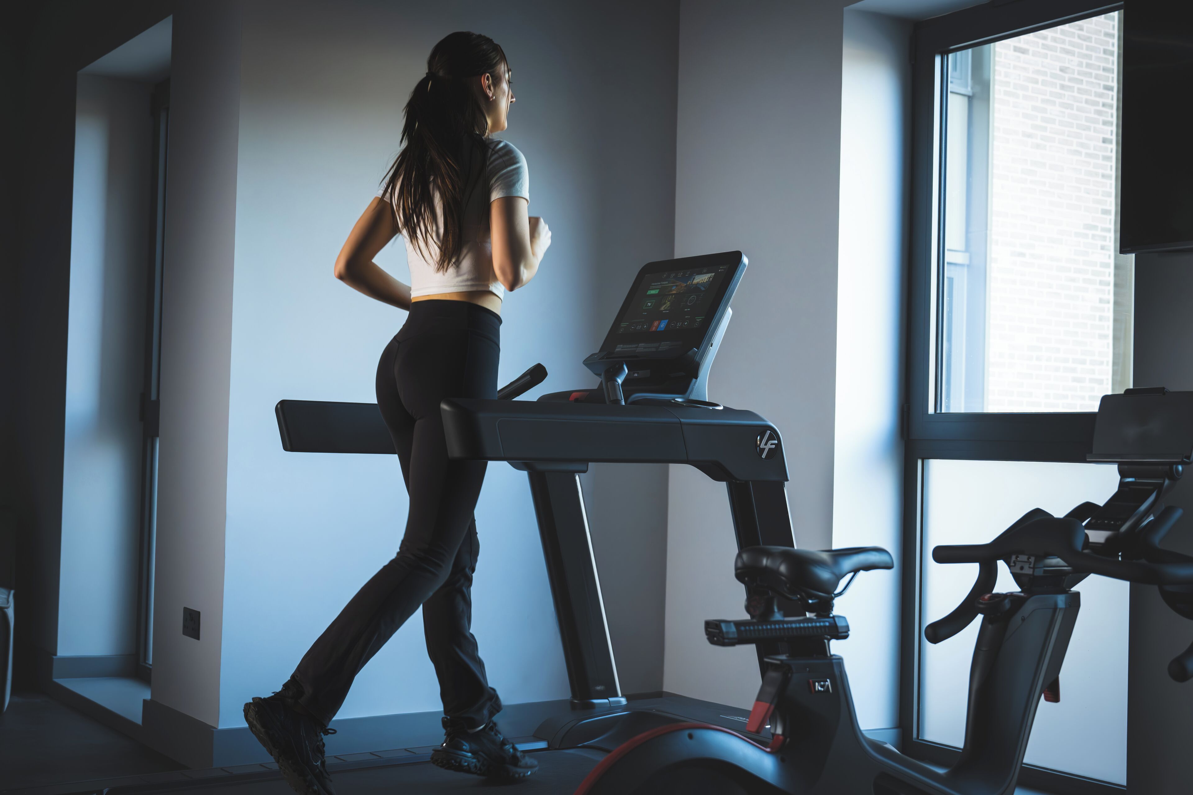 A woman running on a treadmill in a gym with a window and another treadmill in the background.
