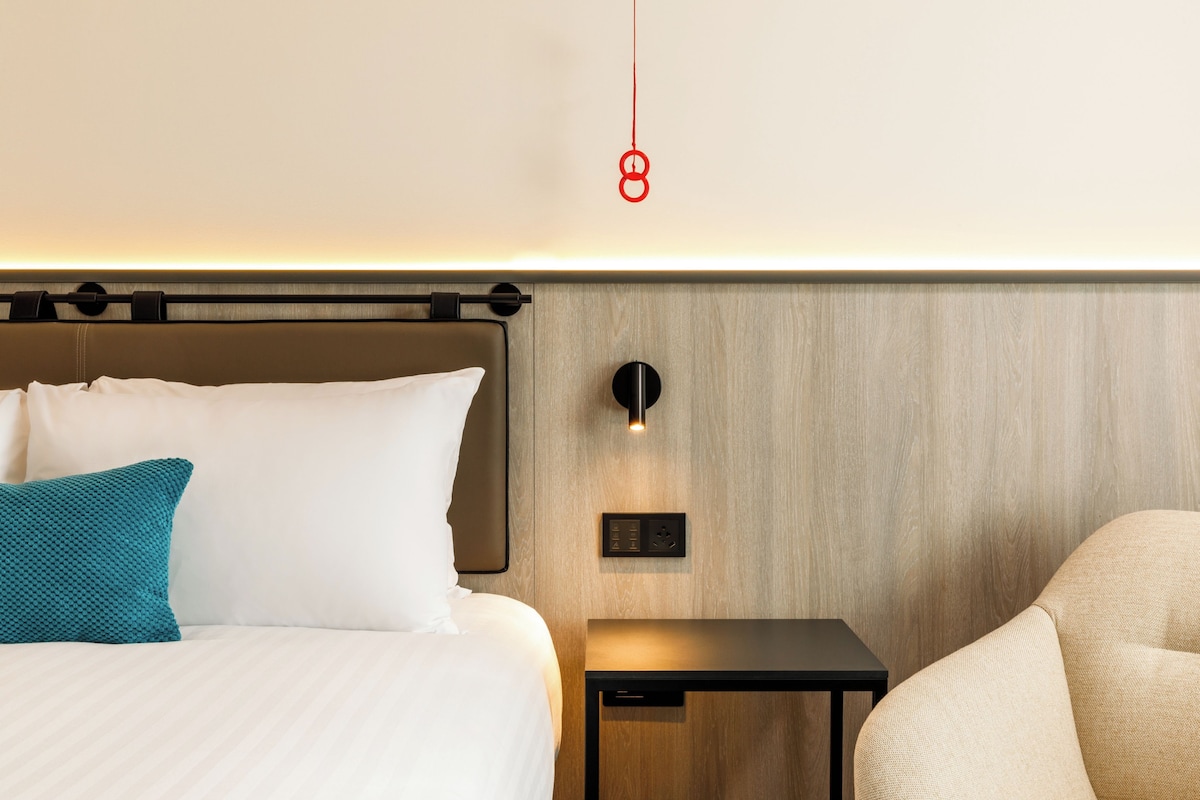 A modern hotel room with a neatly made bed, a bedside table, and a wall-mounted light fixture.