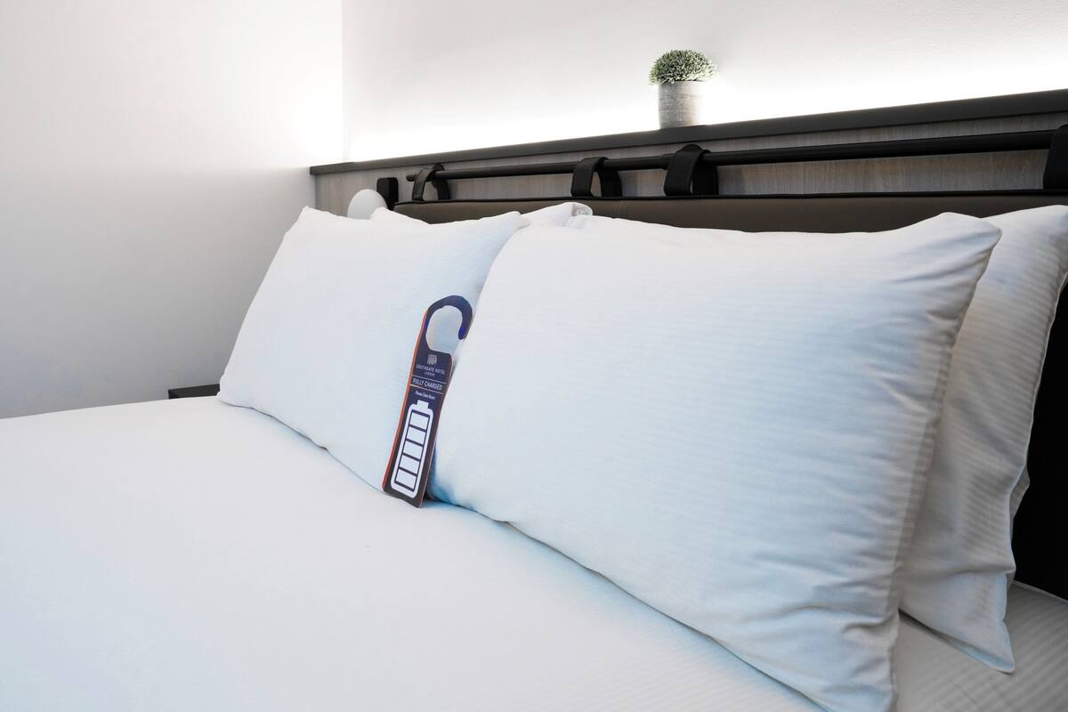 A hotel room with a bed, white bedding, a pillow with a fully charged battery icon, and a small plant on the headboard.