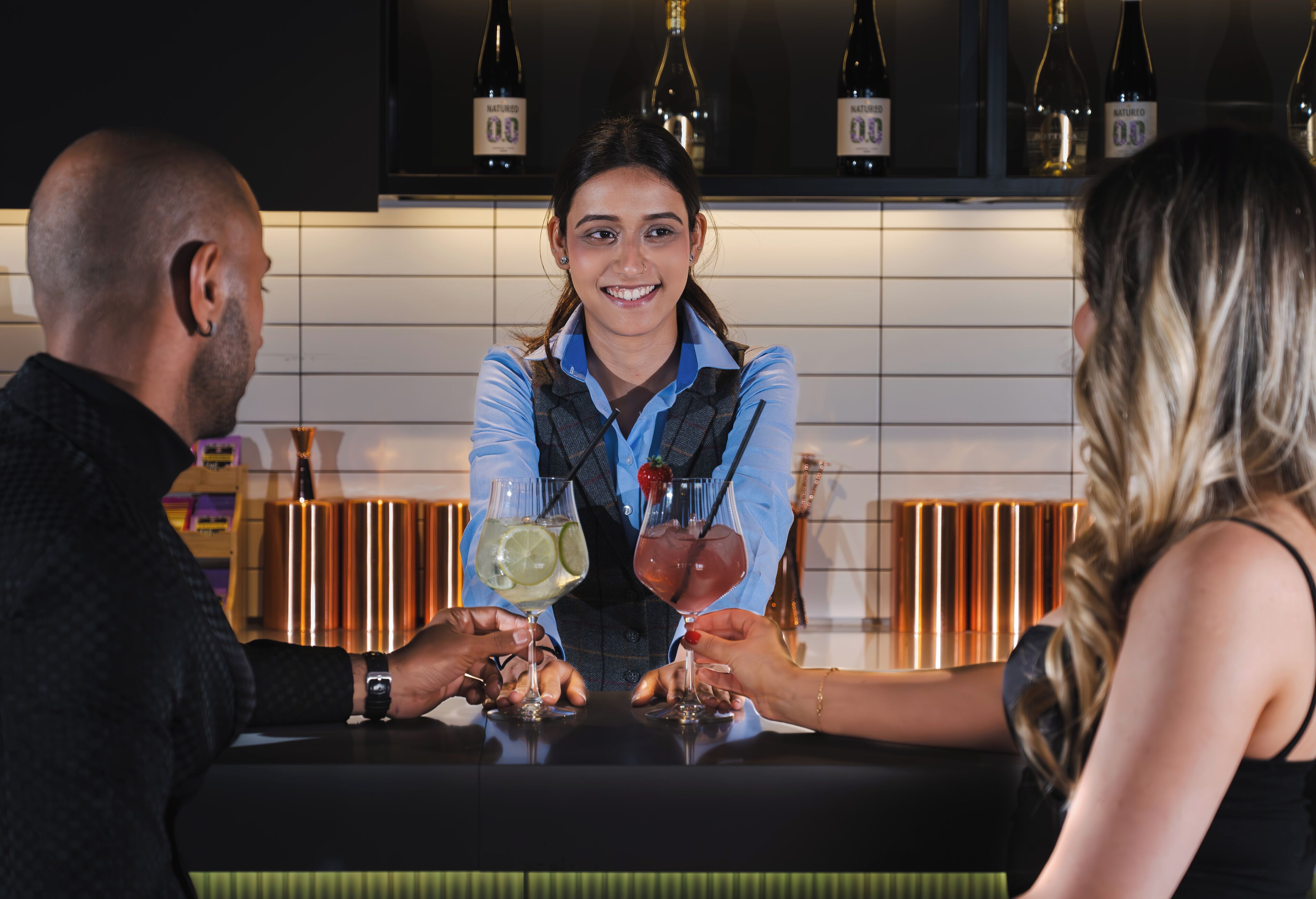 A bar setting with a bartender and two customers. There are bottles of wine on the shelf behind them.