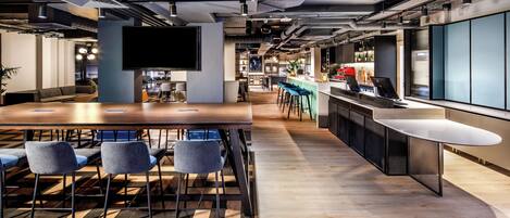 A modern office space with a long wooden conference table, several chairs, a bar area, and a large flat-screen TV mounted on the wall.