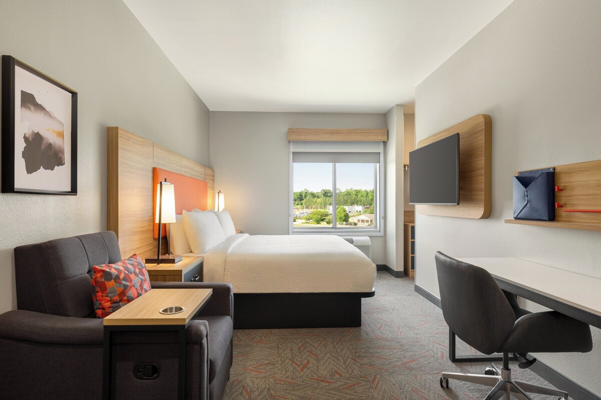 A hotel room with a bed, a desk, a chair, a TV, a lamp, and a window with a view of the outside.