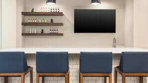 Cozinha moderna com bancada branca, quatro cadeiras azuis, estantes de madeira com garrafas e uma TV preta grande fixada na parede.