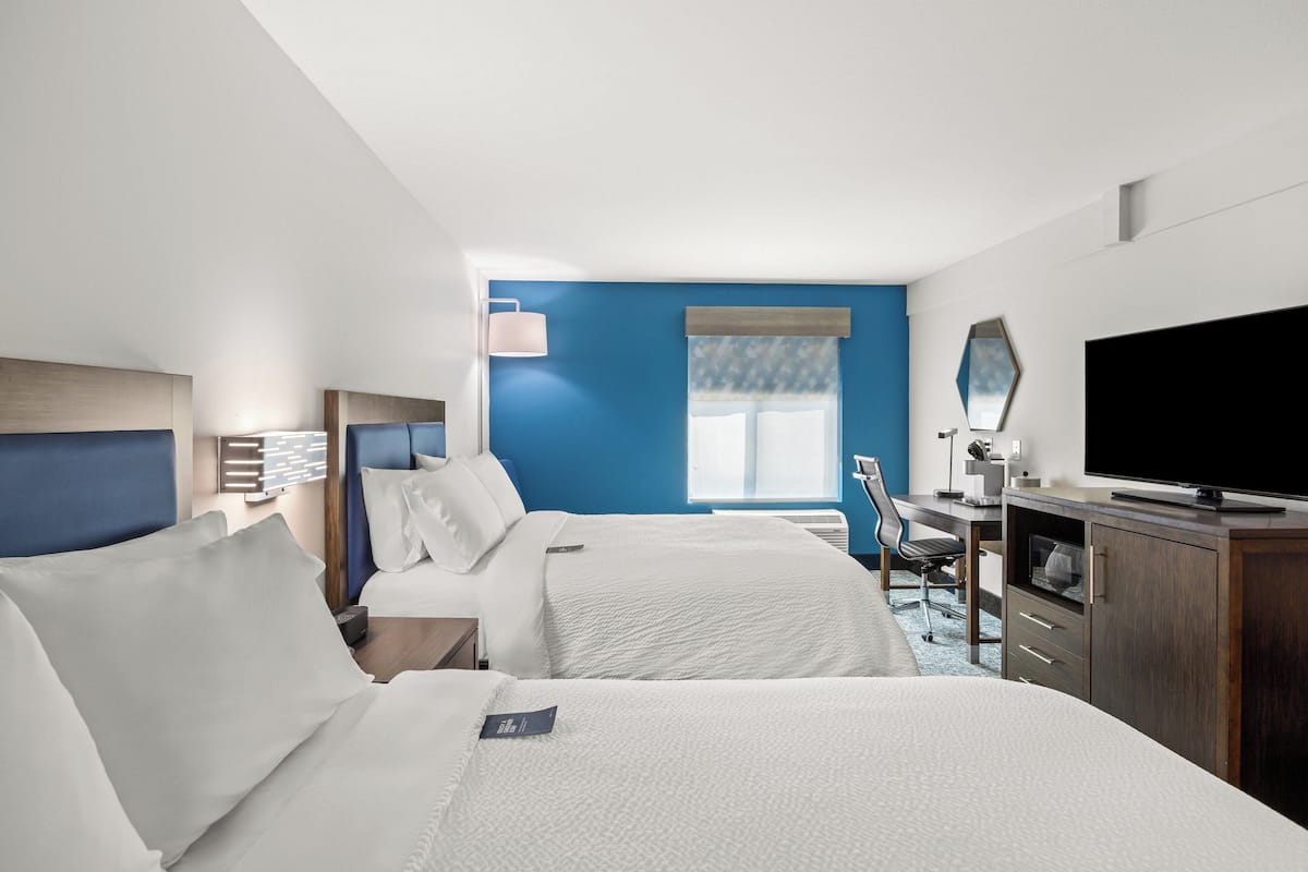 Habitación de hotel con dos camas, un escritorio, televisión y ventana con persianas.