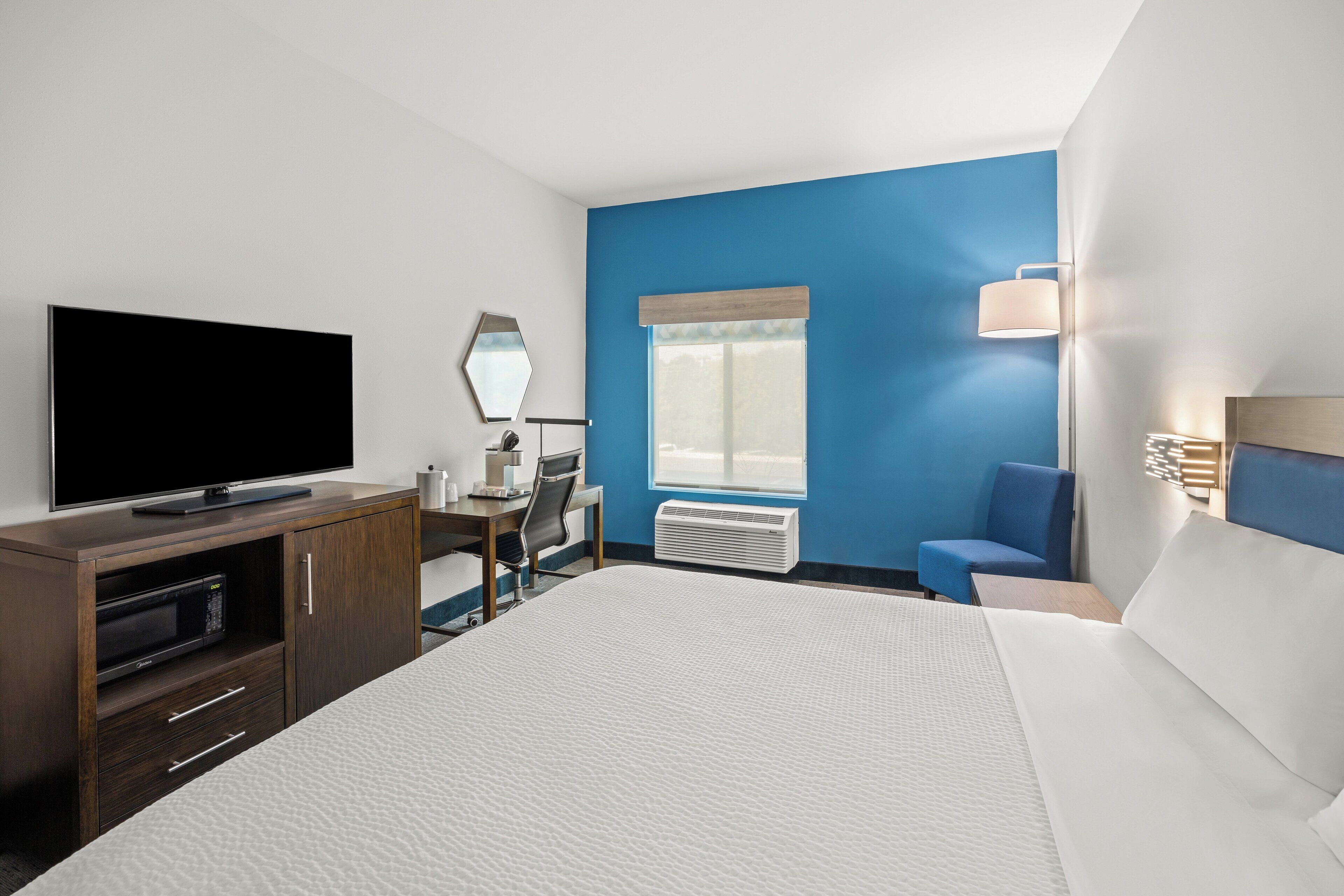 A hotel room with a large bed, a flat-screen TV, a desk with a chair, a window with blinds, and a blue accent wall.
