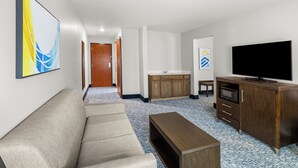 Una habitació d'hotel moderna amb un sofà, un armari de fusta amb televisió de pantalla plana i una zona de cuina amb armaris de fusta, i un quadre abstracte gran a la paret.