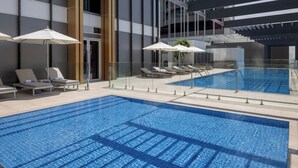 Une piscine intérieure moderne, avec une eau d’un bleu limpide, entourée de chaises longues et de parasols, et dotée d’une rambarde en verre.