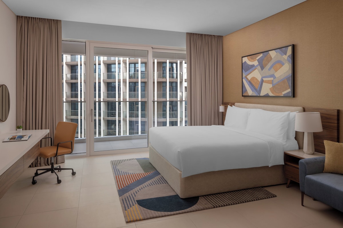 Une chambre d’hôtel avec un lit, un bureau, une chaise, un canapé, une grande fenêtre et un tableau au mur.
