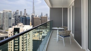 Un balcon doté d’une balustrade en verre, offrant une vue sur un paysage urbain avec de nombreux immeubles de grande hauteur, dont un gratte-ciel imposant. On y trouve une petite table ronde et deux chaises en osier.