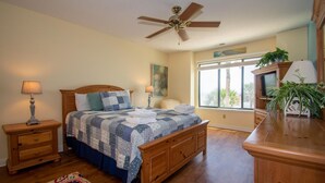 A bedroom with a wooden bed, two bedside tables, a ceiling fan, a window with a view, and a TV on a wooden cabinet. - 646 New Haven Ct (Fripp Island)