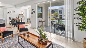 A modern living room with a wooden coffee table, black and pink chairs, and a dining area with a white table and chairs. A large sliding glass door leads to an outdoor balcony with a pool and a view of other buildings. - 3180-207 Resort Pool View Disney Universal Orlando (Kissimmee)