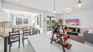 A modern living room with a large white sofa, a coffee table, and a TV mounted on the wall. There are two hanging pendant lights and a wooden coffee table with a Mickey Mouse mug. - 3180-207 Resort Pool View Disney Universal Orlando (Kissimmee)