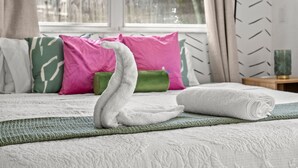 Ein ordentlich bezogenes Bett mit einem schwanenförmigen Handtuch, grün-rosa Kissen und Blick ins Freie durch ein Fenster.