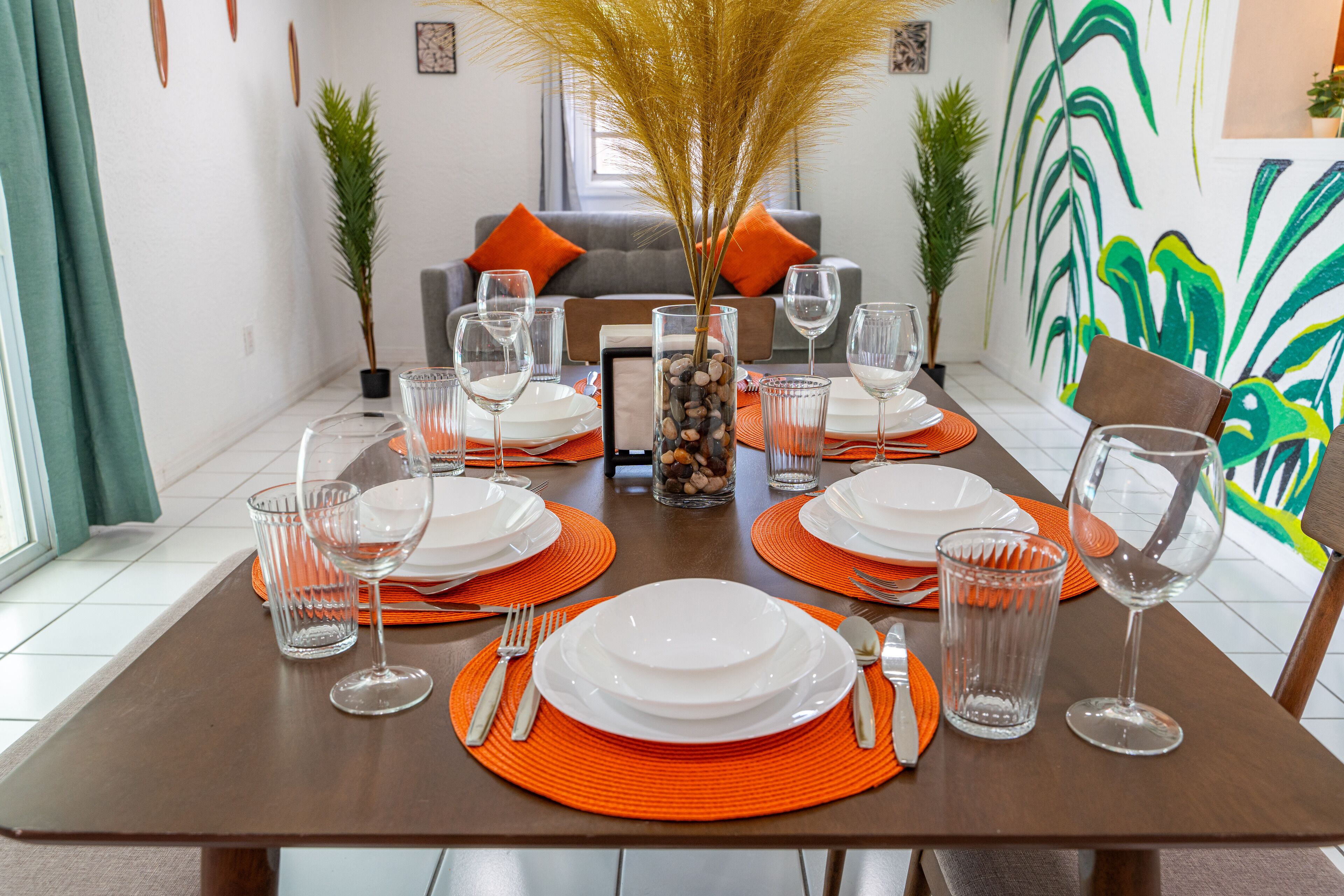 Une table à manger dressée avec de l’assiette blanche, des sets de table orange et de la verrerie transparente.