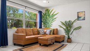 Ein Wohnzimmer mit einem braunen, lederbezogenen Ecksofa, einem kleinen Hocker, einer großen Grünpflanze und einem gerahmten Schild mit der Aufschrift 'Welcome to Florida'.