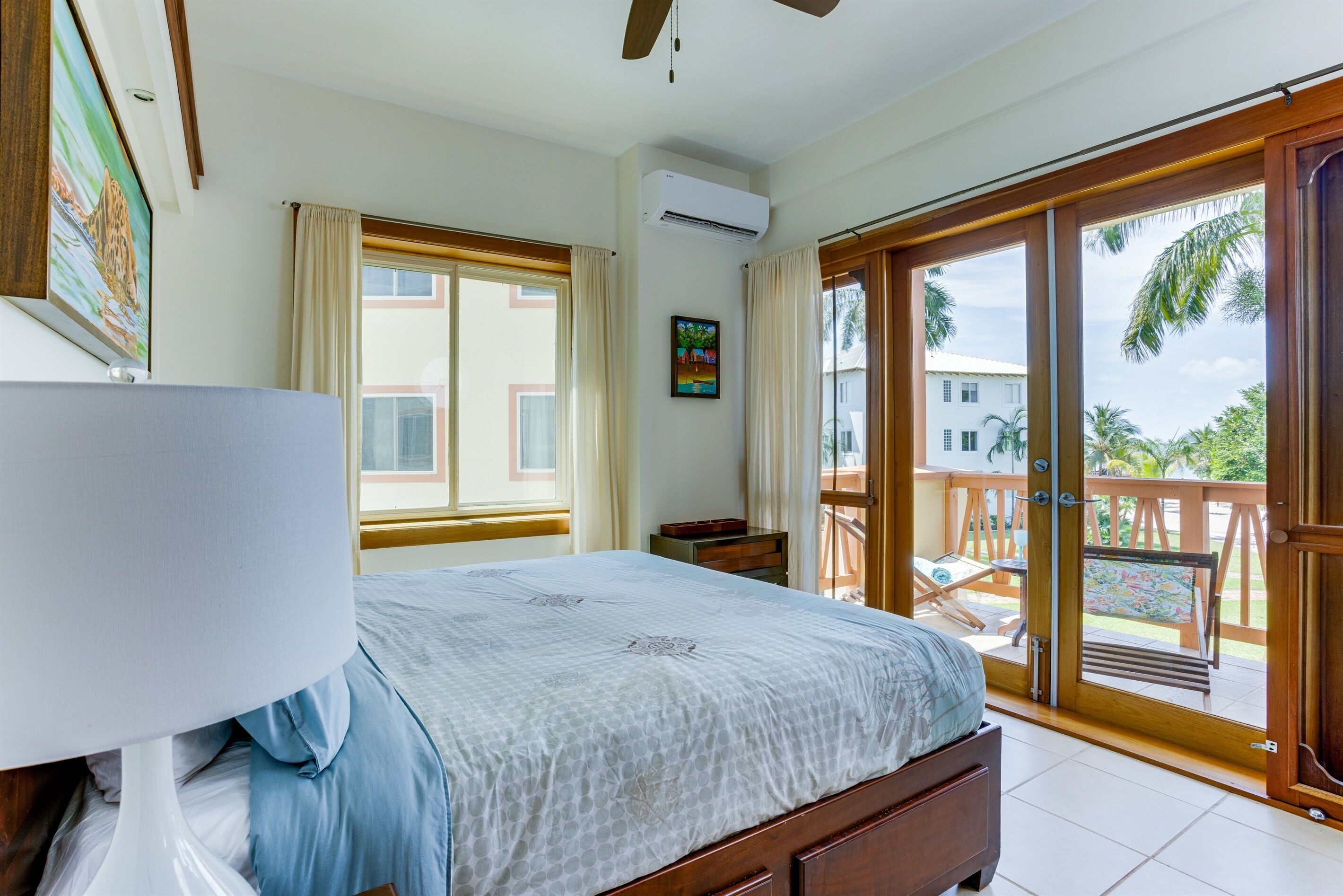 Un dormitorio con cama, mesita de noche, ventana, aire acondicionado y balcón con vista a palmeras.