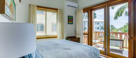 Un dormitorio con cama, mesita de noche, ventana, aire acondicionado y balcón con vista a palmeras.