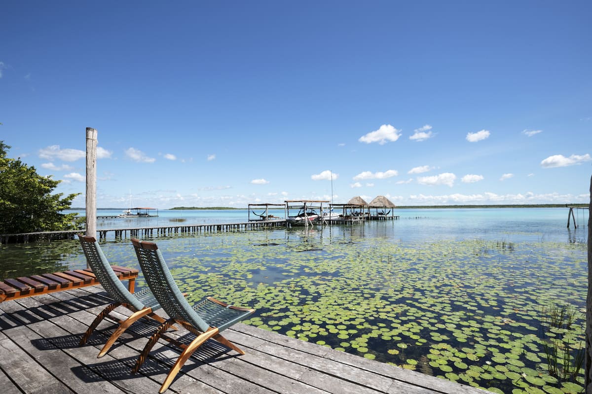 Una terraza de madera con sillones de descanso y vistas a un lago tranquilo con nenúfares, un muelle y bungalows sobre el agua.