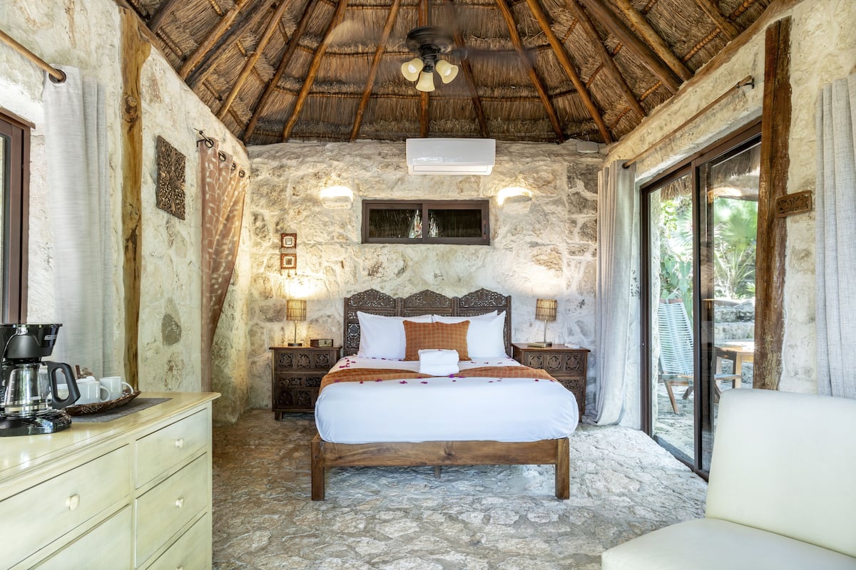 Dormitorio con paredes de piedra, cama de madera, ventilador de techo y ventana con vistas a un jardín.