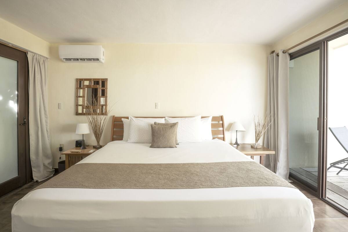 Habitación de hotel con una cama grande, una mesita de noche con lámpara, cabecero de madera, ventana con cortinas y un equipo de aire acondicionado.