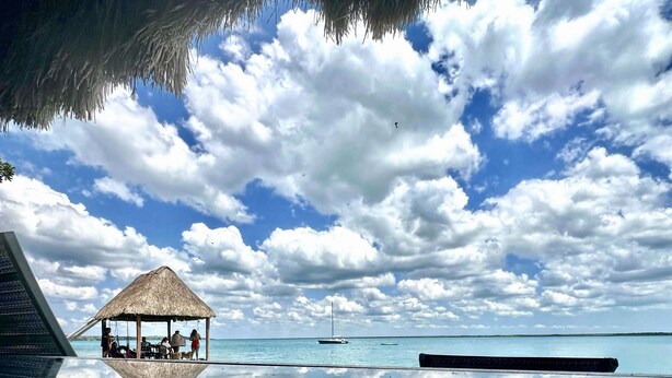A beach with a thatched-roof hut, a wooden deck, and a boat in the distance. - Yayum Bacalar (Bacalar)