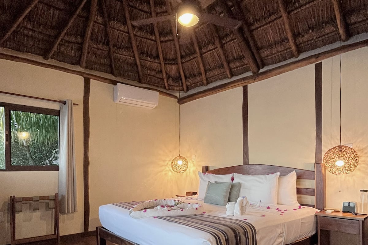 Un dormitorio con techo de madera, una cama con ropa blanca, una mesita de noche y una ventana con cortinas.