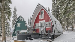 Kaksi jyrkkäkattoista mökkiä, puinen terassi ja lumipeitteinen maa.