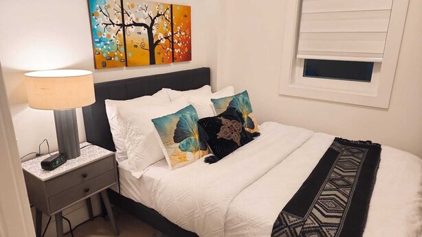 A neatly made bed with white bedding, two decorative pillows, a black throw blanket, a bedside table with a lamp, and a wall-mounted painting.