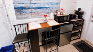 Eine kleine Küchenzeile mit Mikrowelle, Toaster und Kaffeemaschine. An der Wand hängt ein großes abstraktes Gemälde und es steht ein kleiner Tisch mit Stühlen bereit.