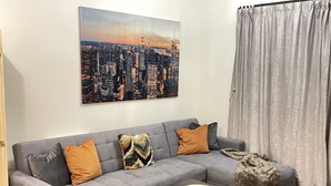 Un salon avec un canapé gris, deux coussins à motifs, une table basse noire sur laquelle repose un bol en bois, et un tableau représentant un paysage urbain accroché au mur.