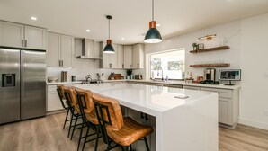A modern kitchen with a large island, stainless steel appliances, wooden bar stools, pendant lighting, and a view to the outdoors.